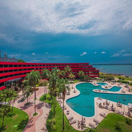 Hotel Royal Tulip Brasilia Alvorada Extérieur photo The photo shows a modern hotel building with a striking red exterior, surrounded by lush greenery. In the foreground, there is a large, creatively shaped swimming pool, with lounge chairs arranged around it. The backdrop features a body of water, pos
